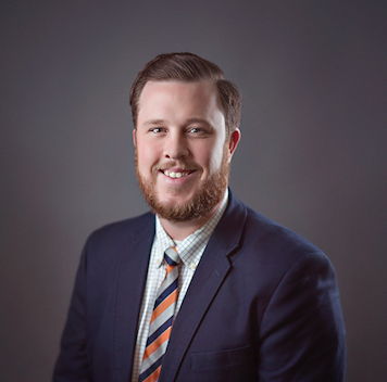 Professional headshot of Dustin Whitlock. He is a white male, with brown hair and beard. He is wearing a suit and smiling.